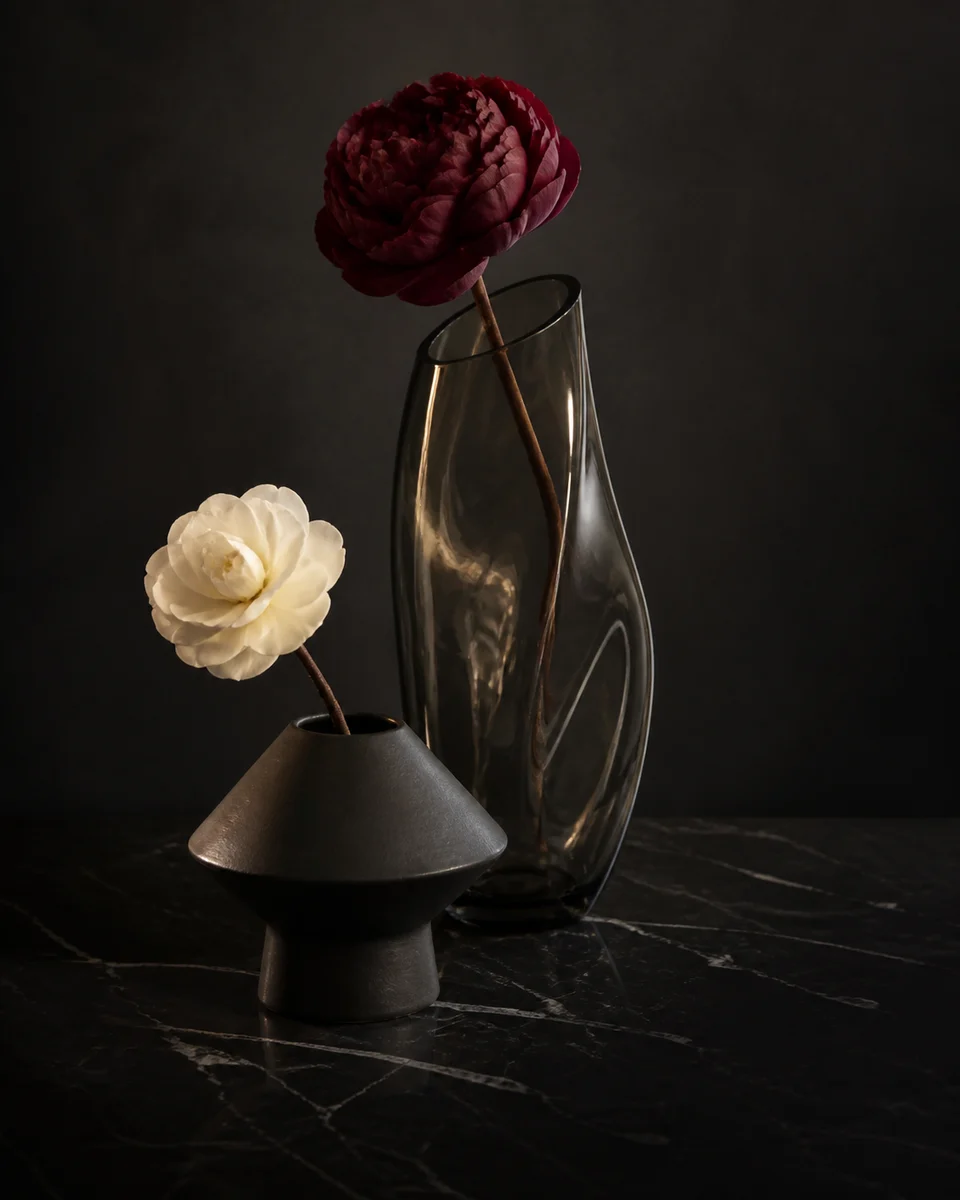 Dos jarrones escultóricos distintos con flores complementarias bajo luz ámbar.